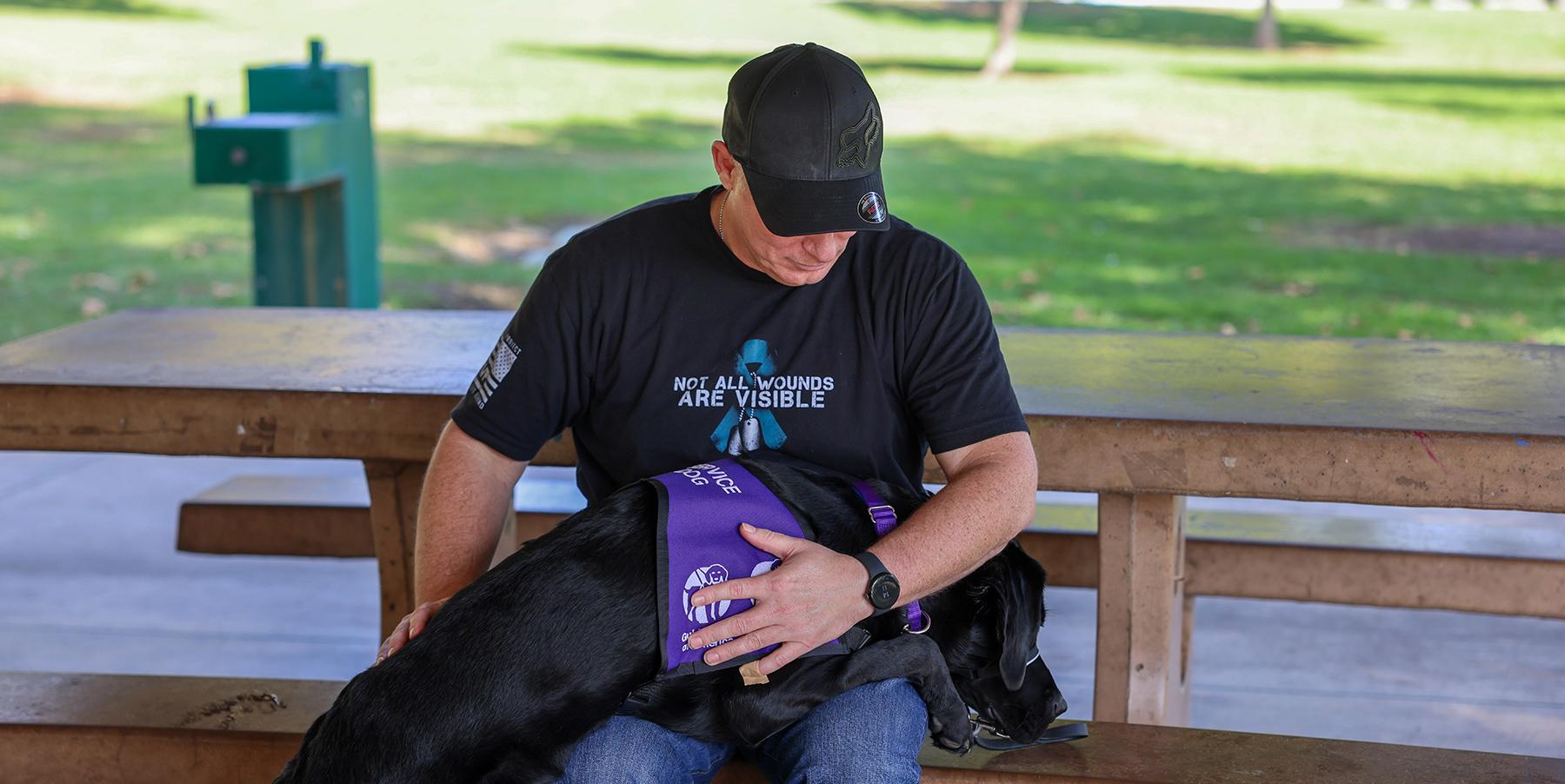 man in black hat and black shirt with black lab in purple vest